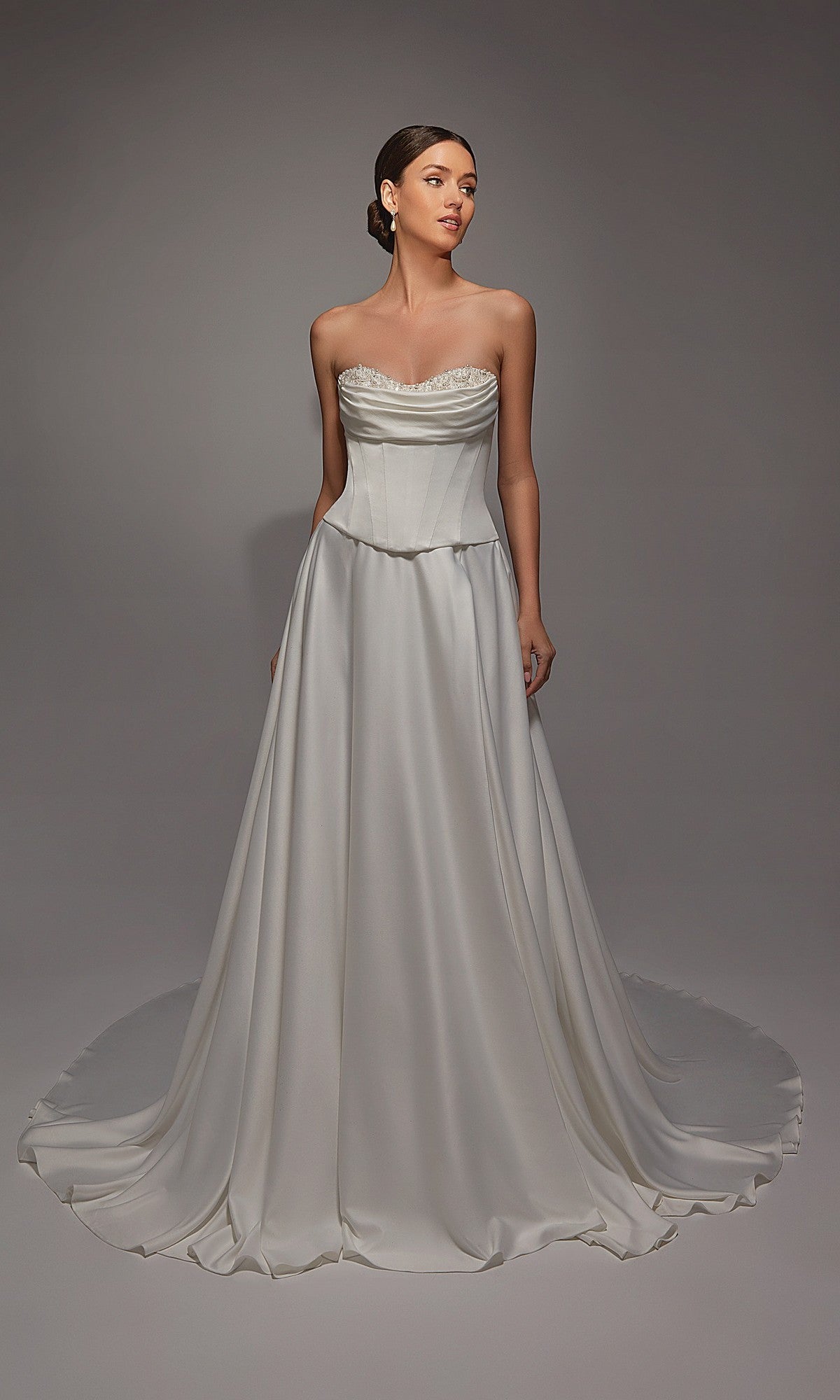 A woman wears the Alyce 7142 long white dress, featuring a satin corset bodice and a full, flowing skirt, standing against a plain gray background.